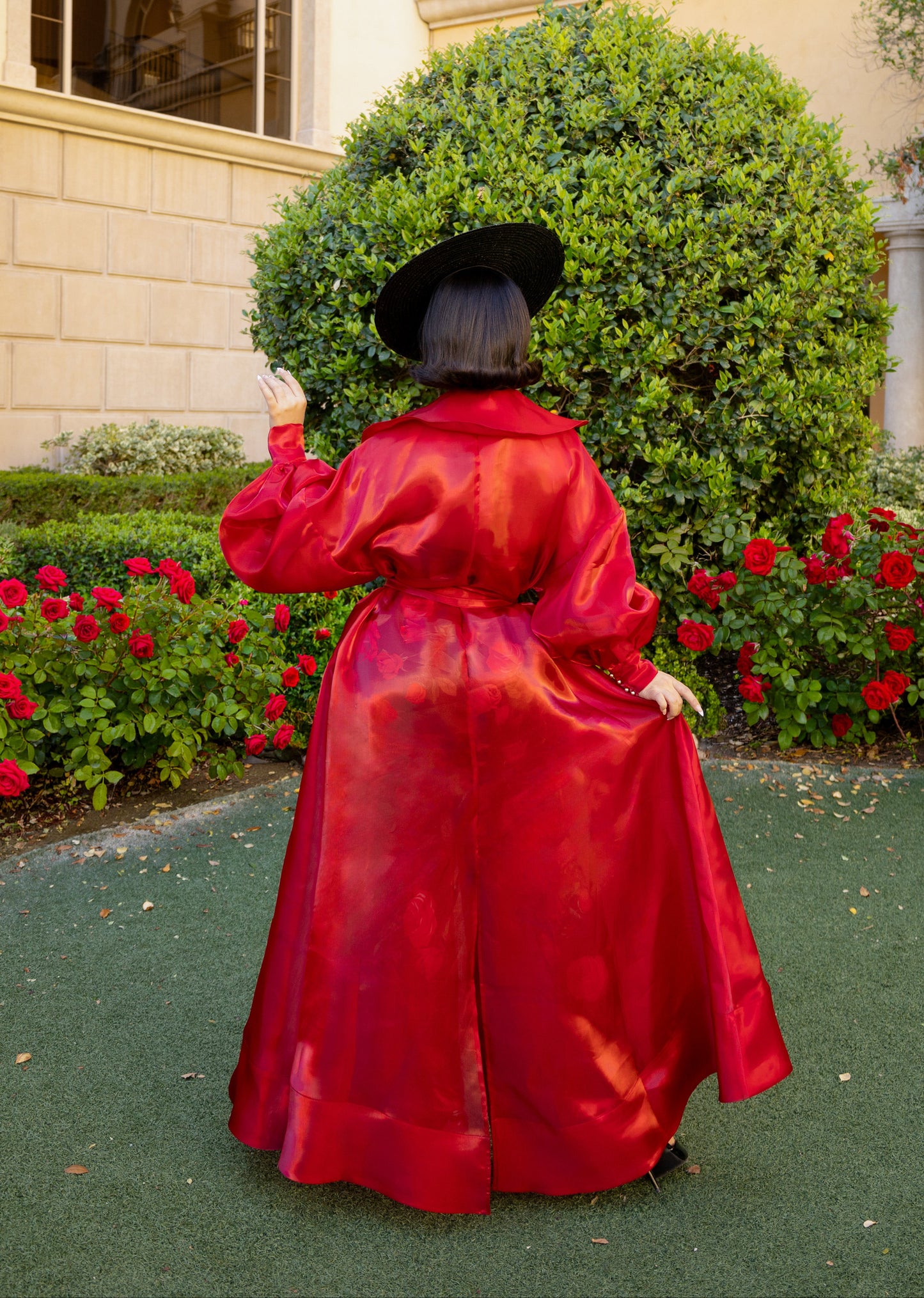 Person in a red overcoat standing in a garden with flowers and a building in the background