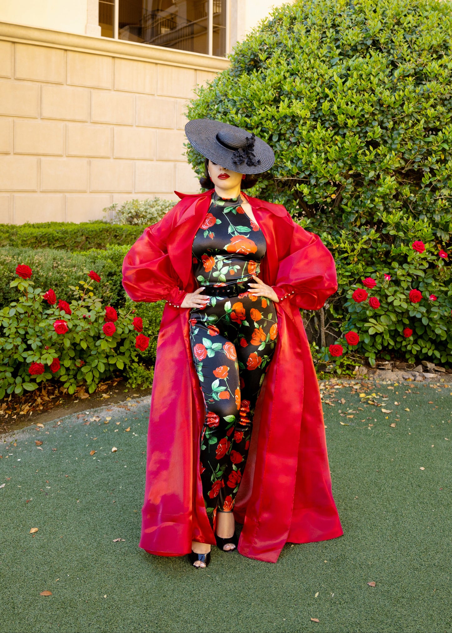 Woman in garden wearing a rose print jumpsuit and red overcoat and a large brim black tilt hat.