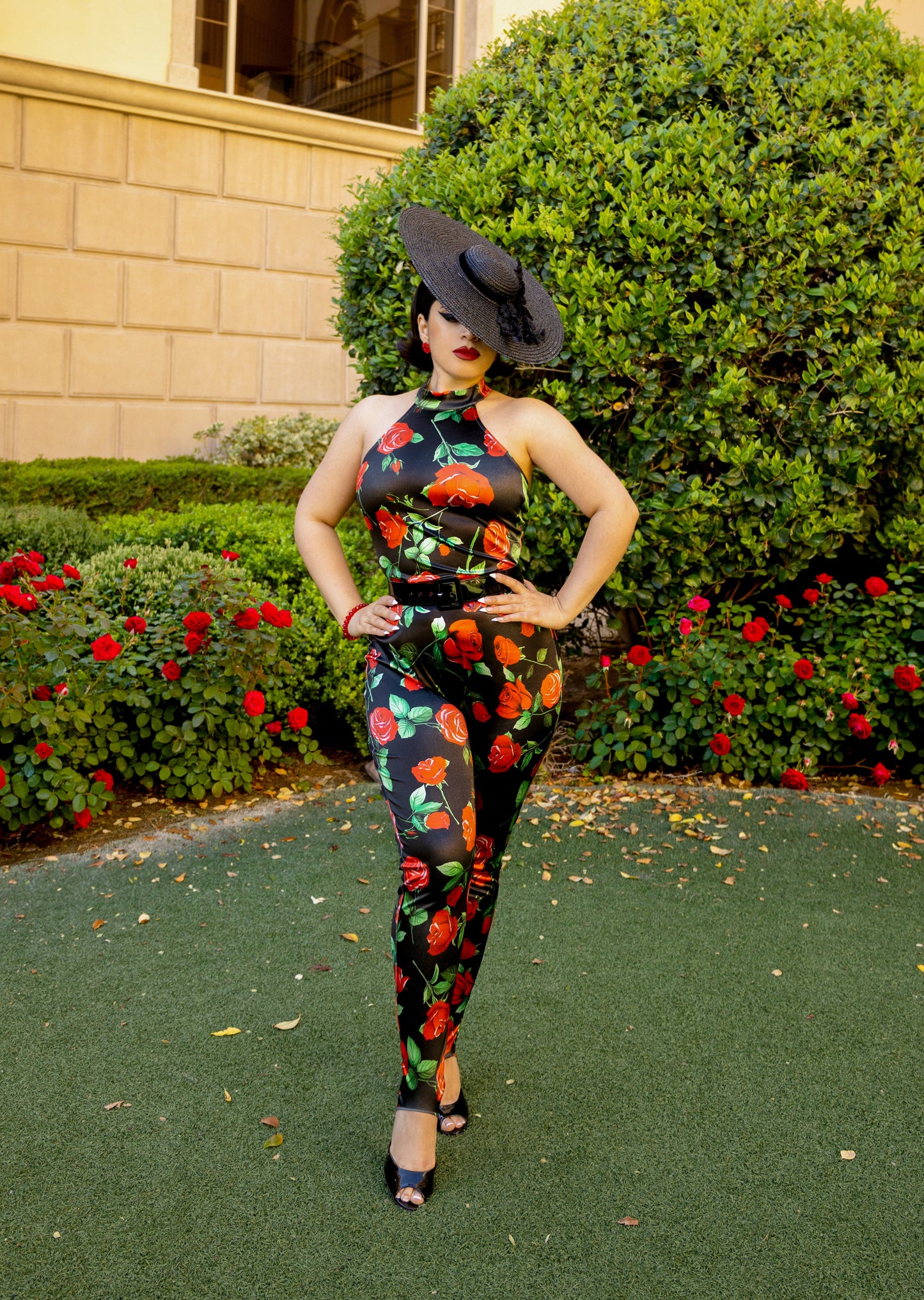 Woman in a floral jumpsuit and hat standing in front of a building with greenery and flowers.