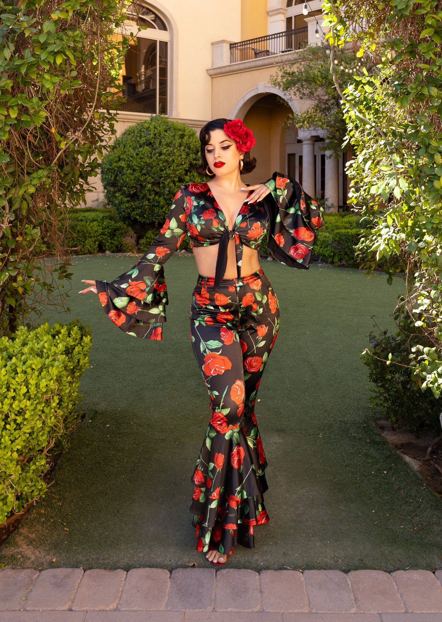 Woman in rose print two-piece bell sleeve top and bell bottom surrounded by greenery.
