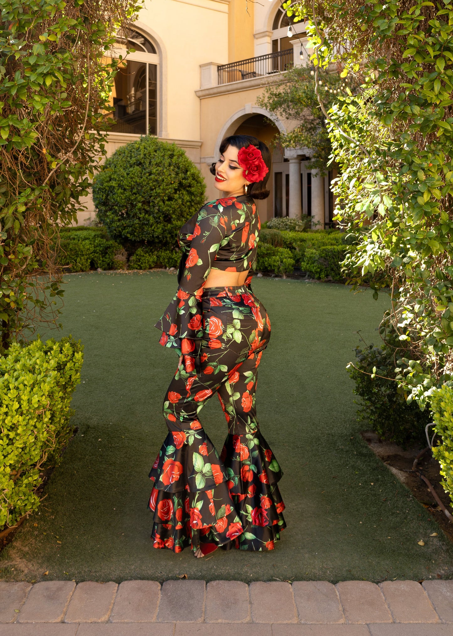 Woman in rose printed bell sleeve top and bell bottom pants surrounded by greenery.