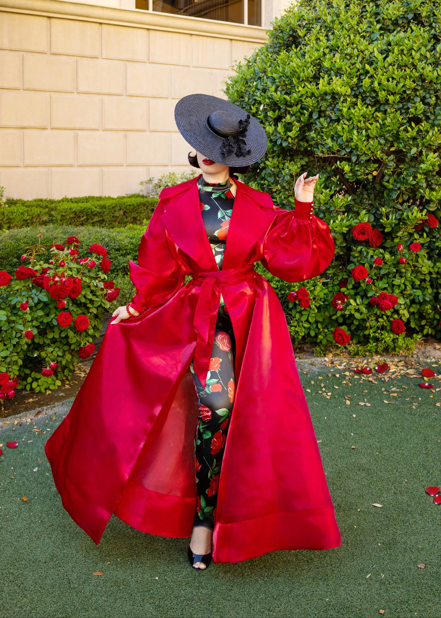 Person wearing a red coat and black hat standing in front of a building with greenery and flowers.