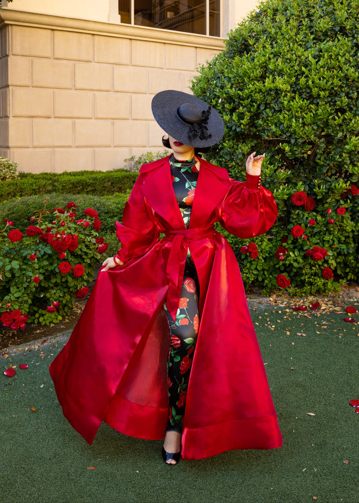 Person wearing a red coat and black hat standing in front of a building with greenery and flowers.