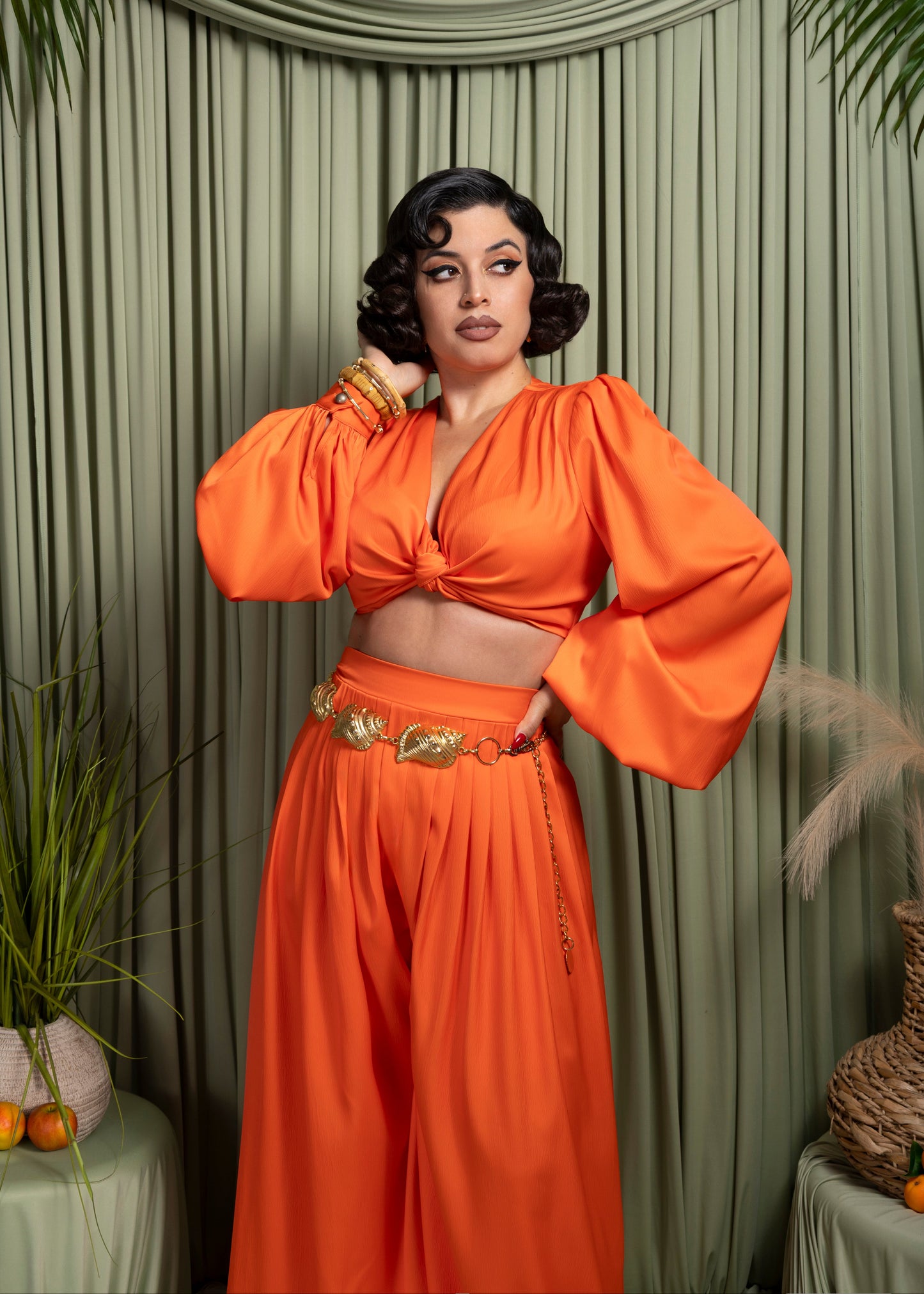 Woman in an orange outfit posing against a green curtain with decorative elements.